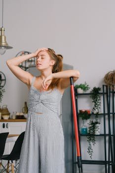 Sleepy young woman in jumpsuit with mop and closed eyes touching head in light house